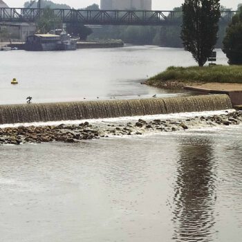Das Bild zeigt das Wehr in der Weser in Hameln. Im Hintergrund sind zwei Schiffe sowie eine Eisenbahnbrücke.